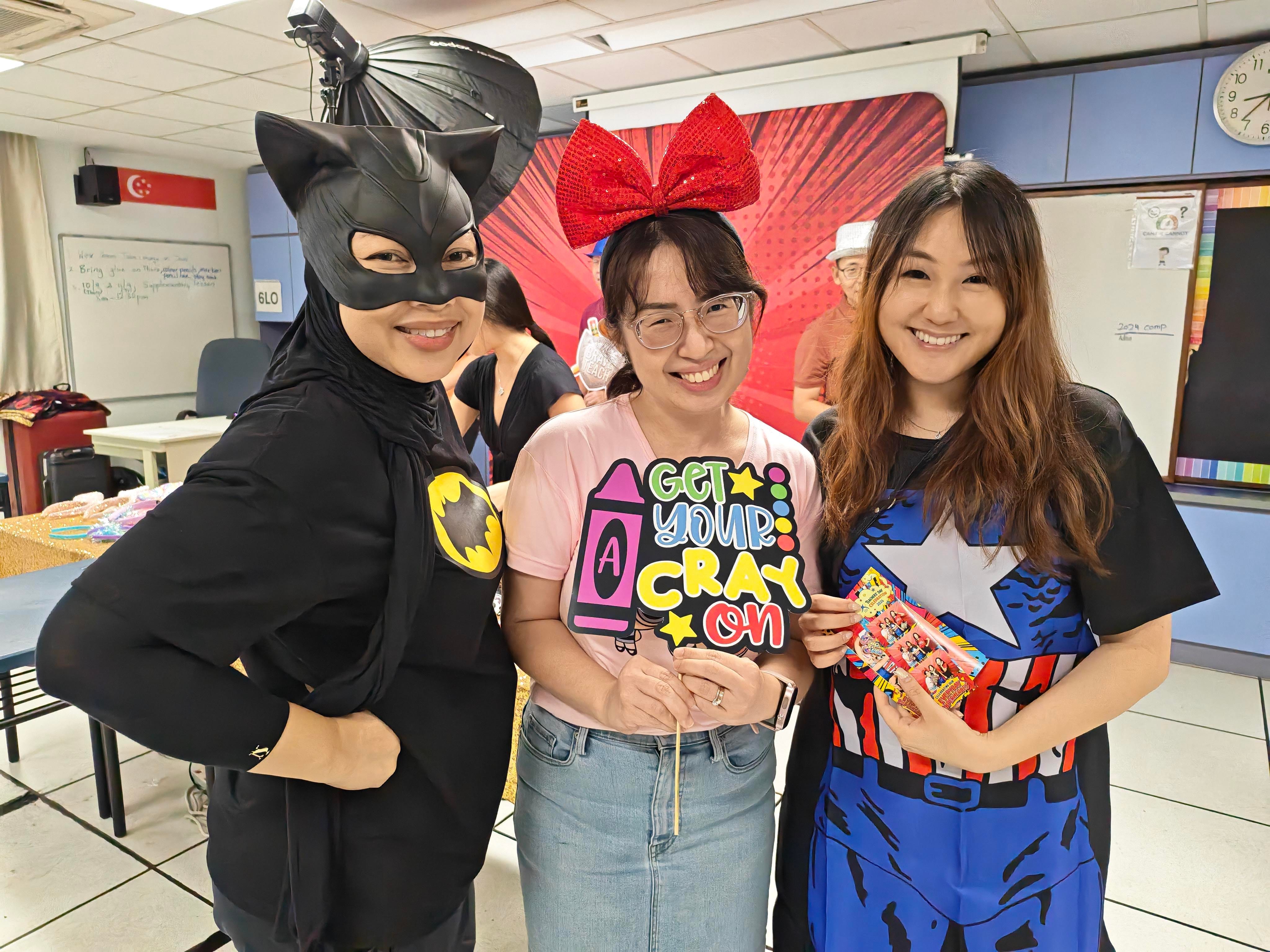 Three smiling teachers pose at a photo booth in a classroom. Two are dressed in superhero costumes, and one holds a colourful "Get Your Crayon" prop while wearing a red bow headband. A comic-style backdrop is set up behind them.